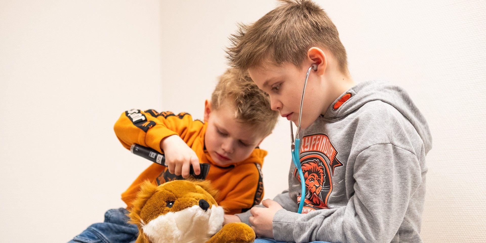 Zwei Kinder untersuchen spielerisch ein Stofftier mit Stethoskop und Ohrenspiegel.