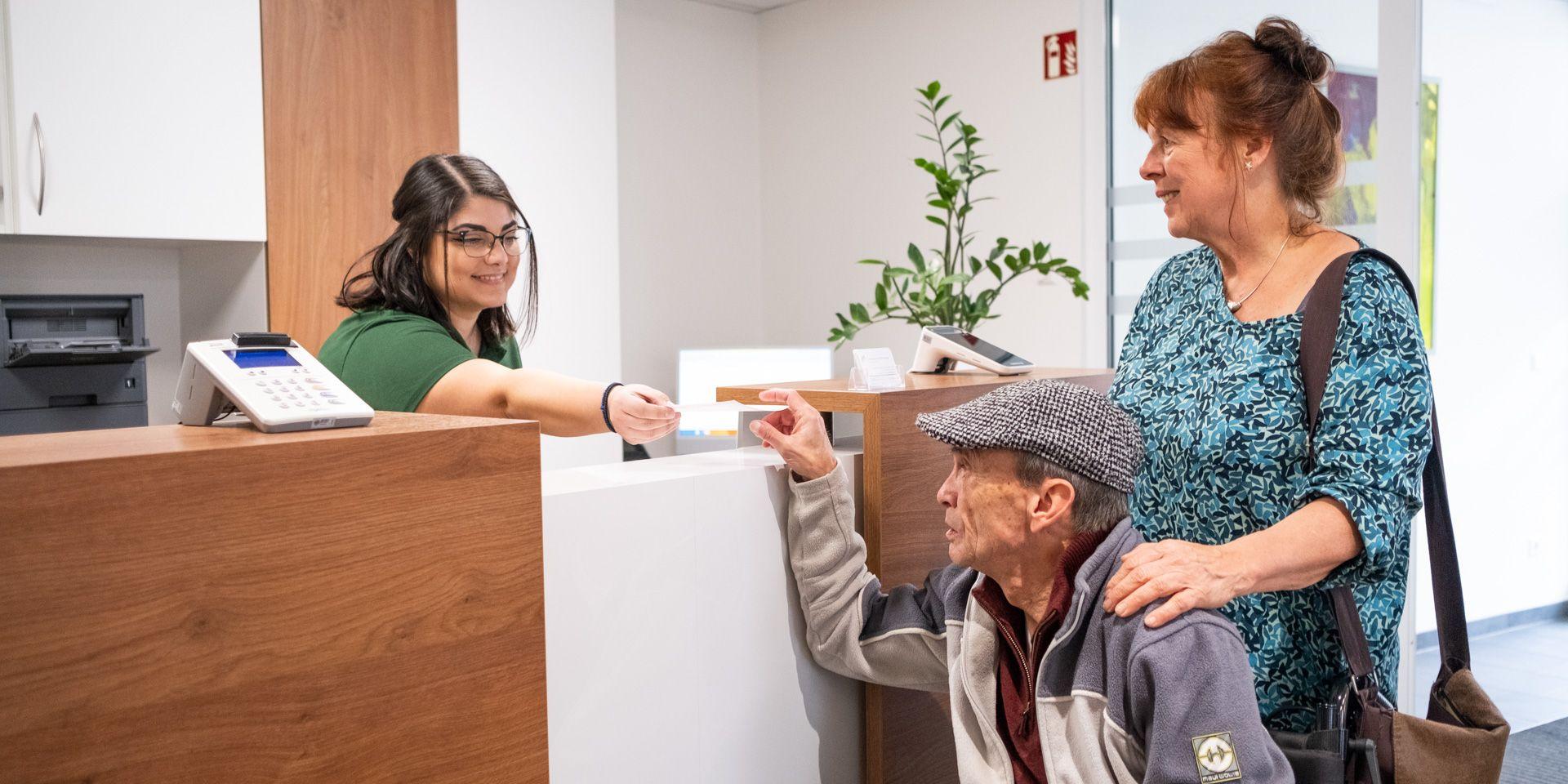 Arzthelferin an der Anmeldung gibt einem Patienten im Rollstuhl seine Versichertenkarte zurück, begleitet von einer Frau.
