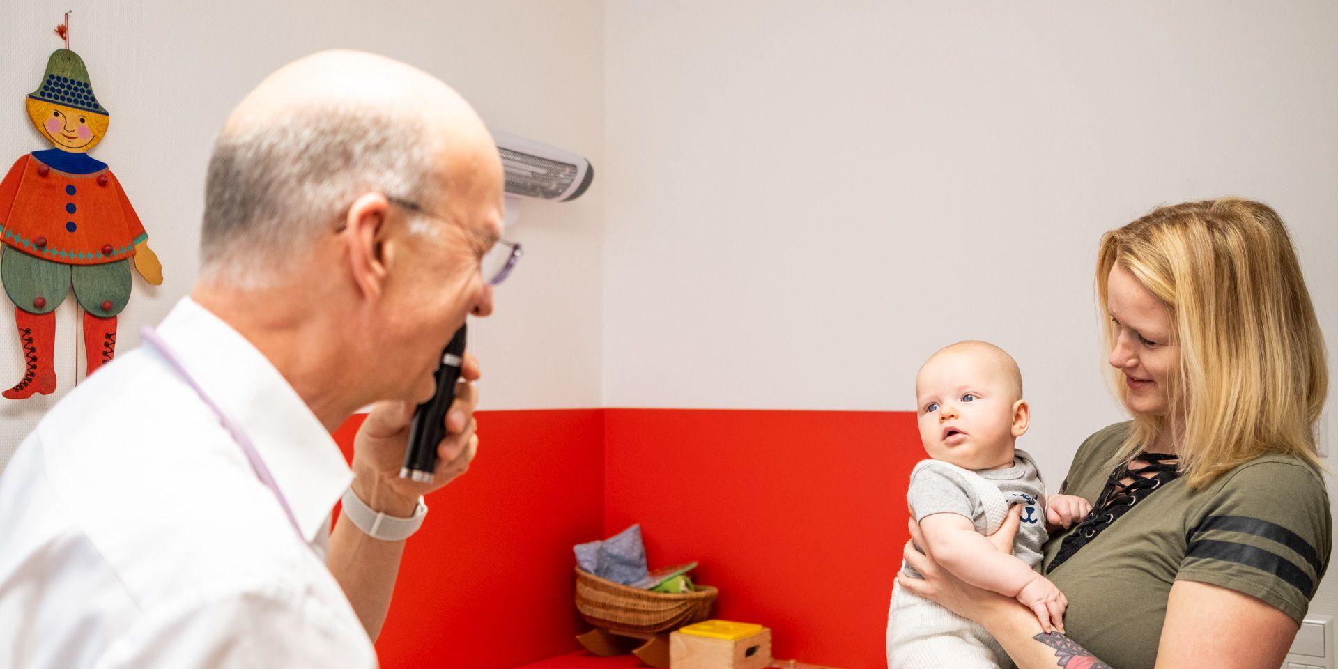 Arzt Joachim Fischer lenkt mit einer kleinen Lampe die Aufmerksamkeit eines Babys, das auf dem Arm der Mutter sitzt.