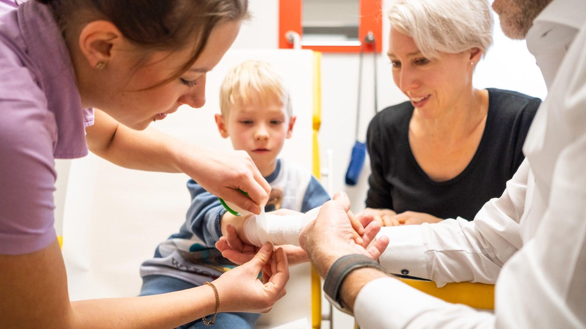 Arzthelferin legt einem Jungen einen Verband am Arm an, Arzt hält die Hand des Kindes